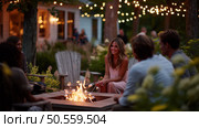 Friends enjoy an evening by the firepit under string lights in a cozy backyard setting during summer. Стоковое AI-изображение, AI-художник Сергей Толмачёв / Фотобанк Лори