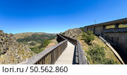 A wooden walkway leading to a dam, surrounded by rugged terrain under a clear blue sky. Guarda, Portugal. Стоковое фото, агентство Ingram Publishing / Фотобанк Лори