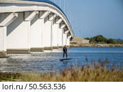 A lone hydrofoil surfer glides beneath a massive white bridge, blending urban architecture with watersport excitement on a calm river. Стоковое фото, агентство Ingram Publishing / Фотобанк Лори