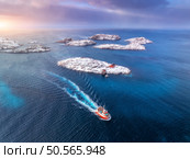 Aerial view of floating fishing boat in blue sea in snowy Henningsvaer village, Lofoten islands, Norway in winter. Top drone view of boat, islands in snow, sea, cloudy sky, town, rorbuer at sunset. Стоковое фото, агентство Ingram Publishing / Фотобанк Лори