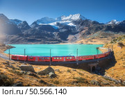 Aerial view of modern red train near azure mountain Bianco lake, snowy rocks and blue sky at sunset in autumn. Bernina Express, Switzerland. Top view of train in Alps, railroad, orange hills in fall. Стоковое фото, агентство Ingram Publishing / Фотобанк Лори