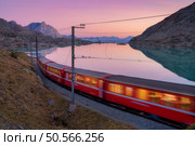 Moving modern red train near azure mountain Bianco lake, snowy rocks and colorful pink sky at sunset in autumn. Bernina Express, Switzerland. Blurred train in Alps, reflection in water in fall at dusk. Стоковое фото, агентство Ingram Publishing / Фотобанк Лори