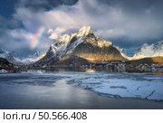 Beautiful reinebringen mountain towers over fishing village Reine in Lofoten islands, Norway, with partially frozen fjord and rainbow in winter at sunset. Dramatic sky over snowy rocks and sea. Travel. Reinebringen mountain dominates the charming fishing village of reine in lofoten islands, norway, with frozen fjord and winter rainbow. Стоковое фото, агентство Ingram Publishing / Фотобанк Лори