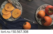 Autumn persimmons on vintage plates, one sliced with a knife, the other whole, on a dark wooden table with a blue napkin and forks. Стоковое фото, агентство Ingram Publishing / Фотобанк Лори
