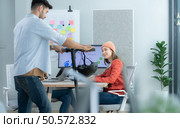 Businessman riding electric scooter in office with colleague in background. Стоковое фото, агентство Ingram Publishing / Фотобанк Лори