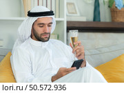 Portrait of a smiling Arabian man holding a glass of champagne and using mobile phone at home. Стоковое фото, агентство Ingram Publishing / Фотобанк Лори
