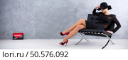 Sexy woman in black dress with hat and red heels sitting on a lounge chair, red-black handbag in the background. Стоковое фото, агентство Ingram Publishing / Фотобанк Лори
