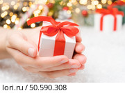 Close up woman hands holding a white gift box with a red ribbon on the background of presents, Christmas tree and golden lights.. Close up woman hands holding a white gift box with a red ribbon on the background of presents, Christmas tree and golden lights. Presents for New Year party. Стоковое фото, агентство Ingram Publishing / Фотобанк Лори