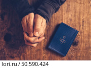 Christian man praying with hands folded and fingers crossed with Holy Bible by his side on wooden desk in church, top view. Стоковое фото, агентство Ingram Publishing / Фотобанк Лори