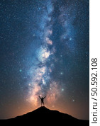 Milky Way and happy man with raised up arms on the mountain peak at starry night. Silhouette of guy, hill, sky with bright stars in summer. Galaxy. Space background. Vertical landscape with milky way. Стоковое фото, агентство Ingram Publishing / Фотобанк Лори