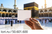 A person holding a blank card with the Kaaba in the background during daylight possibly representing a Hajj or Umrah pilgrimage moment. Стоковое AI-изображение, AI-художник Igor Tichonow / Фотобанк Лори