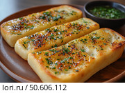 A delicious photo of crispy garlic bread, toasted until golden brown and generously sprinkled with fresh herbs. In the background is a herb and butter sauce in a small dark bowl. Three neat slices are served on a ceramic plate, creating the perfect image of a hearty appetizer or side dish. The image is perfect for food blogs, restaurant menus, bakery ads, and food photography. Стоковое AI-изображение, AI-художник Анна Кривицкая / Фотобанк Лори