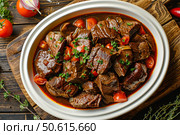 Juicy braised beef stew simmering with tomatoes and herbs in white oval dish. Стоковое AI-изображение, AI-художник Иван Рябоконь / Фотобанк Лори