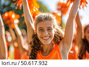 Cheerleader raising orange pom poms and smiling at sporting event. Стоковое AI-изображение, AI-художник Иван Рябоконь / Фотобанк Лори