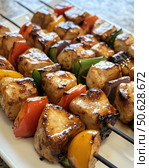 Appetizing kebabs of juicy chicken fillet pieces, fried until golden brown, alternating with pieces of bell peppers of different colors and red onions. The dish is served on a white rectangular plate, which emphasizes the brightness and freshness of the ingredients. Juicy meat shines from the aromatic marinade, and vegetables add color and texture to the dish. An ideal image for culinary sites, recipes, restaurant menus and publications on healthy eating and grilling. Стоковое AI-изображение, AI-художник Анна Кривицкая / Фотобанк Лори