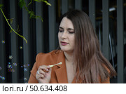 Женщина 38 лет с длинными каштановыми волосами в длинном коричневом платье сидит на стуле на летней веранде с деревянным забором и надула мыльные пузыри и они летают рядом с ней. Стоковое фото, фотограф Светлана Русских / Фотобанк Лори