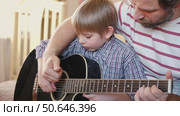 Handsome dad teaches his cute son to play the guitar sitting on sofa in the living room at home. Happy Father's Day. Father teaching his child to play guitar. Kid Learning music skill. Art education. Стоковое видео, видеограф Марина Демидюк / Фотобанк Лори