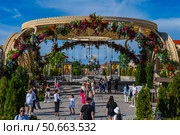 The photo was taken in the cultural and historical complex "New Chersonesus" in Sevastopol. In the foreground is an arched structure decorated with fresh flowers with hanging lamps, forming the entrance to the ceremonial site. Inside are icons, marble pedestals, a landscaped area and many visitors, including children and adults. In the distance, along the axis of the composition, is a majestic temple with a golden dome, symbolizing the spiritual axis of space. The atmosphere is bright, festive, with elements of traditional Orthodox culture and modern landscape design. Редакционное фото, фотограф Анна Кривицкая / Фотобанк Лори