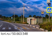 A photograph of a rural bus stop against the backdrop of vineyards in Crimea. A paved road with markings, a pedestrian crossing and road signs leads along a field. The stop is equipped with a canopy and a concrete shelter, on which the word "Shelter" is painted in red. A surveillance camera and electric poles highlight the modern infrastructure. The sky is covered with thick thunderclouds, creating dramatic lighting. The scene conveys the atmosphere of a rural area, removed from the bustle of the city. Стоковое фото, фотограф Анна Кривицкая / Фотобанк Лори