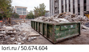 Urban construction site with large green dumpster filled with concrete rubble beside a partially demolished building, surrounded by fencing and debris under overcast sky. Стоковое AI-изображение, AI-художник Катерина Бондарец / Фотобанк Лори