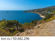 A magnificent view of the Crimean coast. From above, a panorama of the curved coastline, covered with sparse vegetation, rocks and junipers, opens up. In the distance, a small town or village with white houses spread out along the slope of a hill is visible. The Black Sea is deep blue, calm, with slight glare from the sun. On the horizon, closer to the center of the frame, an old sea platform or pier is noticeable. This place conveys an atmosphere of privacy, tranquility and southern warmth. Стоковое фото, фотограф Анна Кривицкая / Фотобанк Лори