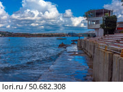 A picturesque view of Sevastopol Bay with a concrete embankment and the steps of an amphitheater facing the sea. Along the shore there is a white building with a terrace, and in the distance you can see ships and cranes of shipyards. The weather is sunny but cloudy, creating a striking contrast between the blue sky, sea and architecture. On the horizon you can see the cityscape of Sevastopol with its historical and industrial sites. Стоковое фото, фотограф Анна Кривицкая / Фотобанк Лори