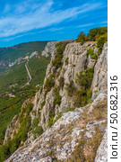 An impressive rocky cliff in the Crimean mountains overlooking a serpentine road that stretches along wooded slopes. In the foreground is a textured cliff surface covered in dry grass and moss. In the distance are mountain ranges, partly covered in greenery, under a bright blue sky with light clouds. Epic and picturesque landscape is perfect for themes of active recreation, mountaineering, hiking and natural power. Стоковое фото, фотограф Анна Кривицкая / Фотобанк Лори