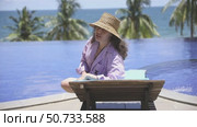 Young woman sits on a lounge chair by a pool at eco beach resort hotel, enjoying the ocean view and palm trees and talking something. Relaxation and tropical vacation vibes. Summer concept. Стоковое видео, видеограф Ирина Михайличенко / Фотобанк Лори