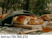 Freshly baked challah bread with honey and floral arrangement on outdoor picnic setting. Стоковое AI-изображение, AI-художник Мария Фадеева / Фотобанк Лори