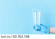 Human hand wear blue nitrile disposable glove holding raised up face mask above light blue background. Safety first concept. Стоковое фото, фотограф Акимова Надежда / Фотобанк Лори