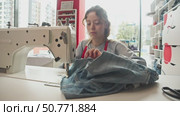 Woman is crafting jeans by sewing them on industrial machine. Measuring tape is draped around her neck as she carefully guides fabric. Стоковое видео, видеограф Strunov David / Фотобанк Лори
