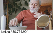 Senior Man shaving his face. Old male shaves beard and mustache with shaver. Elderly gray haired man looking at the mirror, applies shaving cream foam and using razor. Skin hygiene. Daily body care. Стоковое видео, видеограф Марина Демидюк / Фотобанк Лори