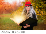 Halloween a woman with a book of magic spells in a witch's hat is conjuring, performing witchcraft against the background of an autumn forest. Стоковое фото, фотограф Лазаренко Светлана / Фотобанк Лори
