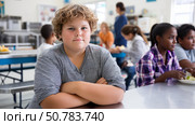 A child sitting alone in a school cafeteria, with others surrounding them, visibly over. Стоковое AI-изображение, AI-художник ALLA YURTAYEVA / Фотобанк Лори