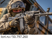 Российский военнослужащий в полной боевой экипировке и защитном шлеме прицеливается из автомата на фоне военной техники в зоне боевых действий. Стоковое AI-изображение, AI-художник ArtVladZ / Фотобанк Лори