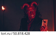 Devilish businessman wearing elegant suit, capturing ironic selfie with smartphone in dimly lit room, red horns visible. Стоковое AI-изображение, AI-художник Данил Руденко / Фотобанк Лори
