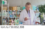 Confident professional senior pharmacist in white coat working behind drugstore counter, inputting purchase orders into computer system, managing incoming supplies and ensuring stock availability. Стоковое видео, видеограф Яков Филимонов / Фотобанк Лори