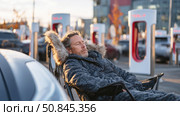 Man sleeping in winter coat beside electric car at charging station. Стоковое AI-изображение, AI-художник Катерина Бондарец / Фотобанк Лори