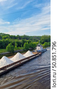 A barge with a cargo of white rubble is moving along the Volga River, which is being dragged by a small tugboat with a Russian flag. Green trees and sparse woodlands are visible on the shore. Стоковое фото, фотограф Анна Чижова / Фотобанк Лори