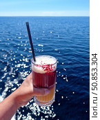 The hand holds a bright double-layered cocktail with red and orange layers and a black tube against a background of sparkling sea surface. The atmosphere of summer relaxation and freedom. Стоковое фото, фотограф Анна Чижова / Фотобанк Лори