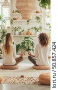 Mindful practice: Two women meditating at home surrounded by plants for a peaceful moment of relaxation. Стоковое AI-изображение, AI-художник Данила Большаков / Фотобанк Лори