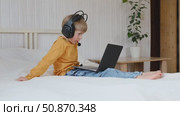 Little smiling 6 years old boy using a laptop with headphones. Child having video call with friends, granny at home in bed. Kid looking at display and waving hand. Childhood leisure activity concept. Стоковое видео, видеограф Марина Демидюк / Фотобанк Лори