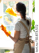 Woman in rubber gloves and apron close-up manually washes window of house with rag cleaner and mop inside interior with home plants on windowsill. Restoring order and cleanliness, cleaning servise. Стоковое фото, фотограф Ольга Симонова / Фотобанк Лори