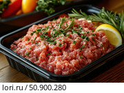 Fresh minced chicken neatly laid out in a black plastic container. Decorated with herbs, rosemary sprigs and a lemon wedge. In the background are vegetables and fresh herbs, creating an appetizing and homely atmosphere. The image is suitable for illustrating food products, a healthy lifestyle, culinary recipes and online stores. Стоковое AI-изображение, AI-художник Анна Кривицкая / Фотобанк Лори