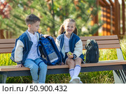Two sibling children with a backpack sit on a bench near the school after classes and talk to each other. Children friendships and relationships of classmates. Стоковое фото, фотограф Ирина Михайличенко / Фотобанк Лори