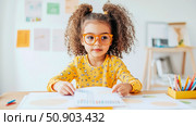 A cute little girl studying at desk, wearing glasses and a bright yellow shirt, focused on her learning material, creating a charming scene. Стоковое AI-изображение, AI-художник Данила Большаков / Фотобанк Лори