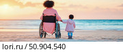 Concept: Intergenerational Support; A woman in a wheelchair and a child holding hands on the beach at sunrise, symbolizing family love and togetherness. Стоковое AI-изображение, AI-художник Данила Большаков / Фотобанк Лори