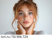 Portrait of a young girl with freckles and expressive eyes, looking straight into the camera. Her face is framed by light red hair, gathered in a light hairstyle, and her hands gently support her chin. Soft natural light and a neutral background emphasize the purity and tenderness of the image. Suitable for themes of youth, natural beauty, skin care, aesthetics and portrait photography. Стоковое AI-изображение, AI-художник Анна Кривицкая / Фотобанк Лори