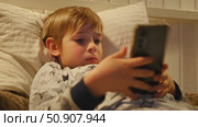Cute kid boy in pajamas lying in bed watching smartphone, smiling. Children's screen time. Child plays games using phone at the evening or night. Social media addiction. Childhood, modern technology. Стоковое видео, видеограф Марина Демидюк / Фотобанк Лори