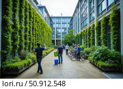 Modern eco friendly office building with vertical green walls and people in courtyard. Concept of sustainable architecture and healthy workplace. Стоковое AI-изображение, AI-художник Павел Муравьев / Фотобанк Лори